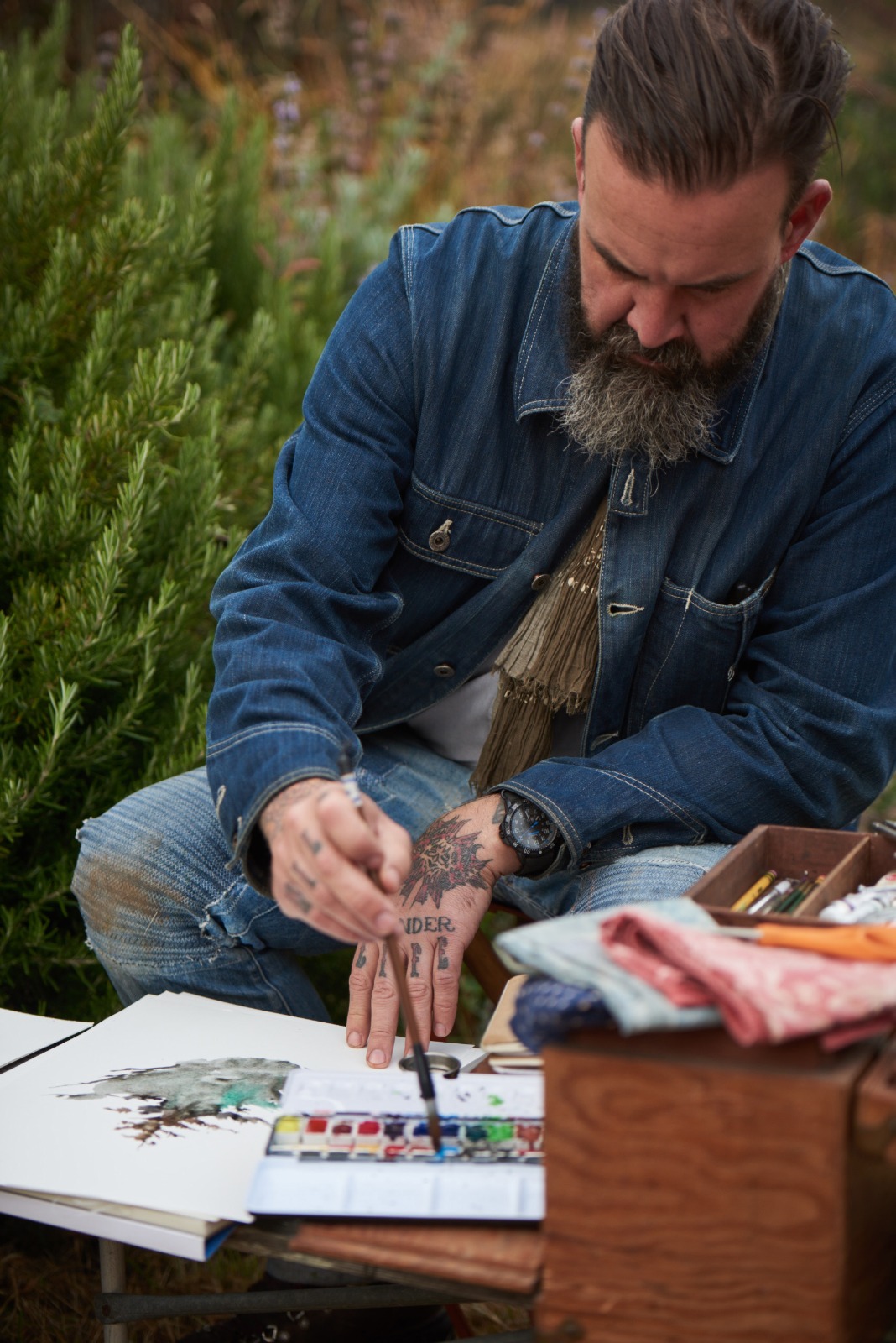 Obi Kaufmann painting a watercolor outdoors, brush in hand, beside a rosemary shrub